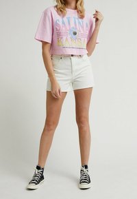 Vaaleanpunainen lyhythihainen t-paita, jossa on "SALINA KANSAS" -grafiikka ja kukkakuosi, yhdistetty valkoisiin farkkushortseihin ja mustiin korkeatossuihin.