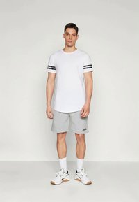 Λευκό T-shirt με μαύρες ρίγες στα μανίκια, συνδυασμένο με ανοιχτό γκρι σορτς. Το μοντέλο φοράει λευκές κάλτσες και sneakers με μπεζ σόλα.