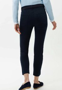 Tmavě modré slim kalhoty s hladkou texturou, s mírně zvýšeným pasem a zadními kapsami. Stylizované s světle modrým svetrem a tmavými botami.