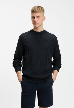 Uomo biondo con capelli corti, indossa un maglione a coste scuro e pantaloncini blu scuro, in piedi con le mani nelle tasche davanti a uno sfondo chiaro e uniforme.