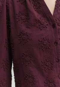 Bordeaux shirt met geborduurde bloemmotieven, voorzien van een v-hals, knoopsluitingen en textuurdetail over het gehele oppervlak.