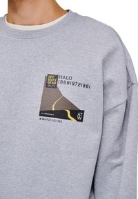Grå sweatshirt med rund hals, med en grafisk patch i svart, gult och brunt som föreställer ett berg och en vägdesign.