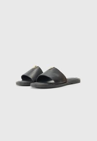 Sandales à glisser en cuir noir avec un design à bout ouvert, semelle plate et un accent décoratif doré à l'avant. Texture lisse et forme minimaliste.