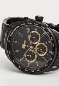 Montre chronographe en acier inoxydable noir avec un cadran noir, des accents dorés, trois sous-cadrans et une échelle tachymétrique autour de la lunette.