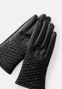 Un par de guantes de cuero negro con un patrón de costura en forma de diamante acolchado en los puños, con dedos lisos, dispuestos sobre una superficie blanca.