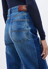 Τζιν ψηλόμεσο, από ανοιχτό έως σκούρο μπλε τζιν ύφασμα με άνετη εφαρμογή, με καφέ δερμάτινο ταμπελάκι στη μέση και δύο πίσω τσέπες.