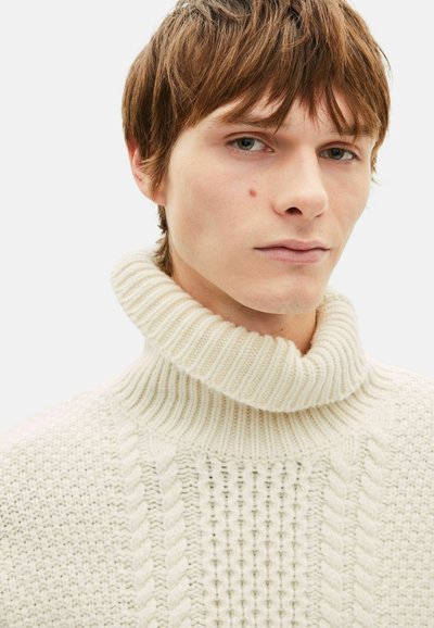 Suéter de punto color crema con cuello alto acanalado, con patrones texturizados, trenzas en la parte frontal y un diseño suave y grueso.