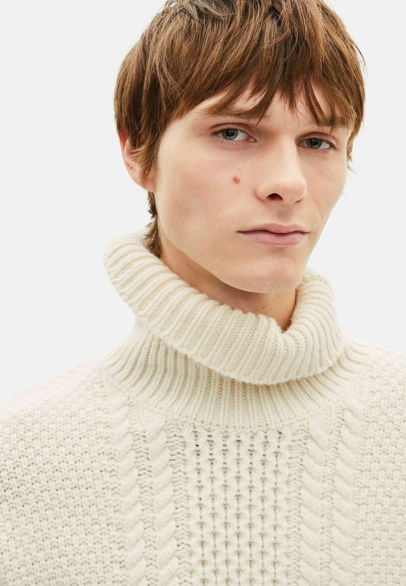 Pull en tricot couleur crème avec un col roulé côtelé, présentant des motifs texturés, des torsades sur le devant et un design doux et épais.