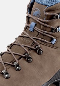 Stivale da trekking in suede marrone con accenti blu chiaro, occhielli metallici e tessuto testurizzato attorno al polsino. Sistema di allacciatura robusto visibile.