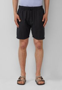 Zwarte, elastische shorts met een relaxte pasvorm, voorzien van een enkele knoopsluiting. Gecombineerd met bruine sandalen met dubbele gespen.