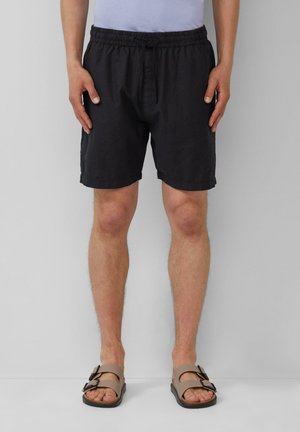 Shorts noirs à taille élastique avec une coupe décontractée, présentant une fermeture à un seul bouton. Associés à des sandales marron avec deux boucles.