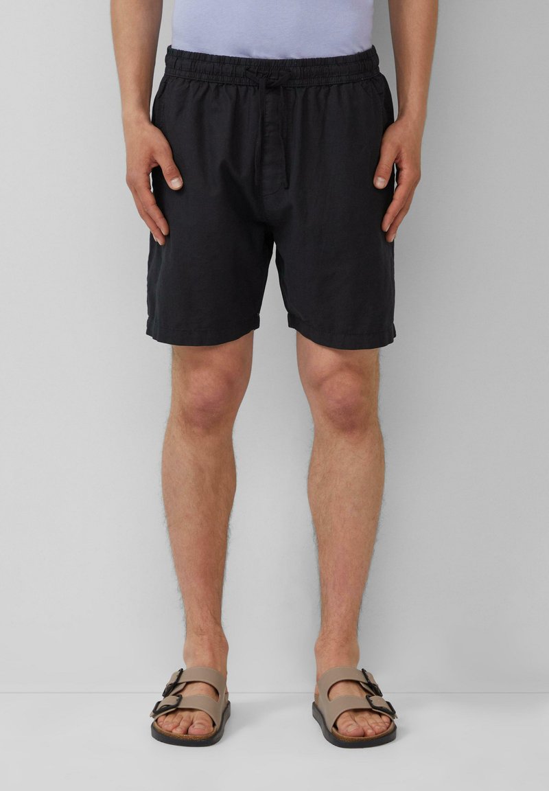 Zwarte, elastische shorts met een relaxte pasvorm, voorzien van een enkele knoopsluiting. Gecombineerd met bruine sandalen met dubbele gespen.