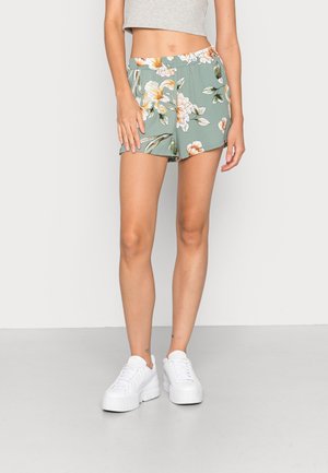 Femme portant un short vert à fleurs, un crop top blanc et des baskets compensées blanches, debout devant un fond uni clair.