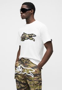 Λευκό βαμβακερό μπλουζάκι με μεγάλο γραφικό σχέδιο παραλλαγής. Συνδυασμένο με πράσινα παντελόνια καμποτάζ με λευκό ζωγραφιστό μοτίβο. Μαύρα γυαλιά ηλίου.