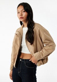 Beżowa kurtka bombowiec z zapięciem na zamek, ściągaczami przy mankietach i bocznymi kieszeniami, połączona z białym topem crop i ciemnoniebieskimi dżinsami.