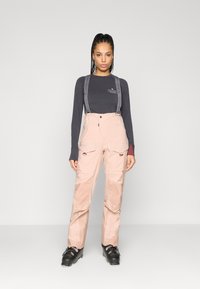 Calças de esqui em rosa claro com suspensórios cinza ajustáveis, múltimos bolsos cargo e textura suave. Camisola de manga longa escura usada por baixo.