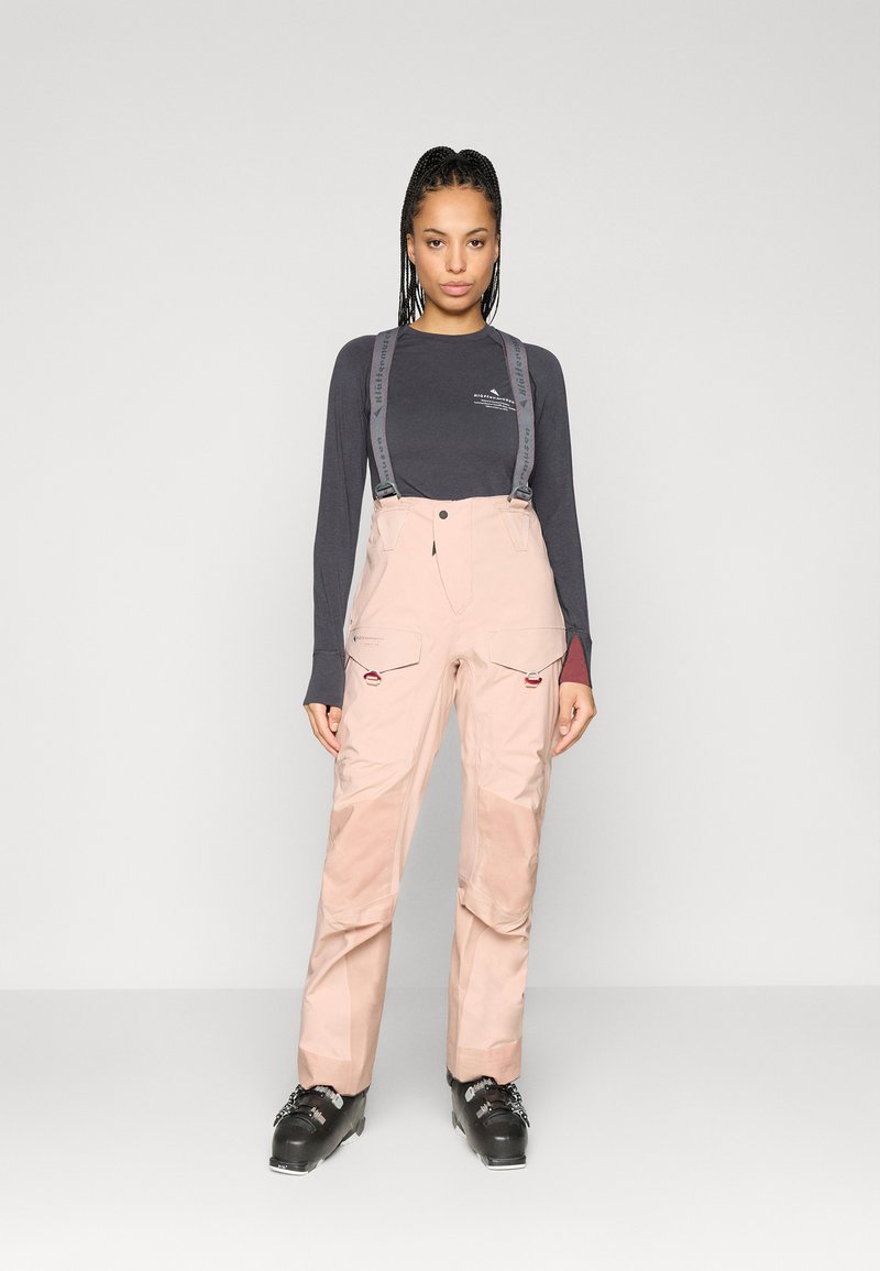 Calças de esqui em rosa claro com suspensórios cinza ajustáveis, múltimos bolsos cargo e textura suave. Camisola de manga longa escura usada por baixo.