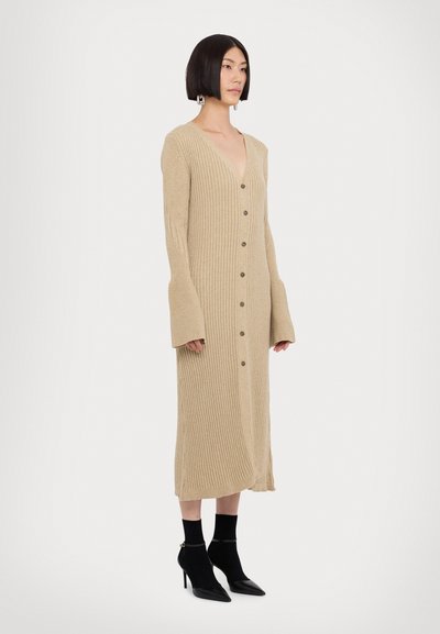 Beige geribbelde gebreide cardigan jurk met lange mouwen, diepe V-hals, knoopsluiting aan de voorkant en zijsplit, gecombineerd met zwarte hoge hakken.