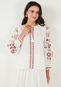 Blouse en lin blanc avec de longues manches bouffantes, ornée de broderies florales rouges et d'un détail de lien à l'avant. Texture douce, coupe décontractée.