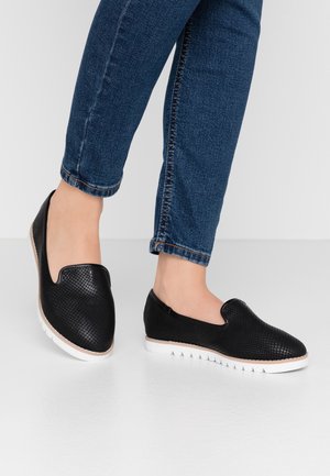 Voeten met zwarte slip-on schoenen met een textuur en witte zolen, gecombineerd met blauwe spijkerbroek, tegen een effen lichte achtergrond.