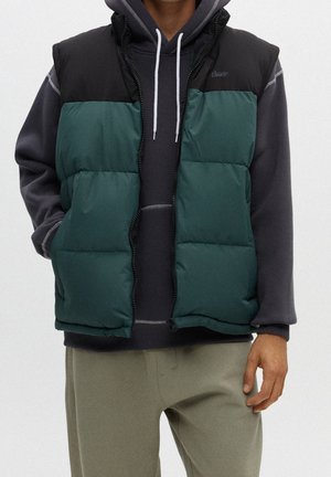Man draagt een groen-zwart donsvest over een donkergrijze hoodie met witte trekkoorden en lichtgrijze broek, staat met één hand in de zak.