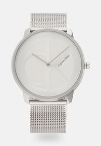 Montre avec bracelet en maille argentée, dotée d'un cadran rond minimaliste avec un fond blanc, un logo en relief et des aiguilles de heures et de minutes élégantes.
