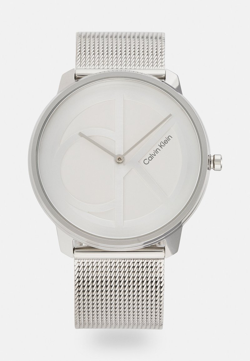 Montre avec bracelet en maille argentée, dotée d'un cadran rond minimaliste avec un fond blanc, un logo en relief et des aiguilles de heures et de minutes élégantes.