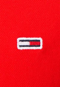 Un tejido rojo exhibe un pequeño logo bordado rectangular con un borde blanco, que presenta Rayas horizontales en azul y rojo.