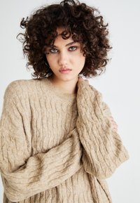 Beige kabelstrikket genser laget av mykt materiale, med rund hals og oversized ermer, som viser et strukturert mønster.