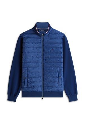 Jachetă albastră matlasată cu fermoar frontal, două buzunare cu capse, manșete și tiv striate și mic logo roșu, alb și albastru pe piept.