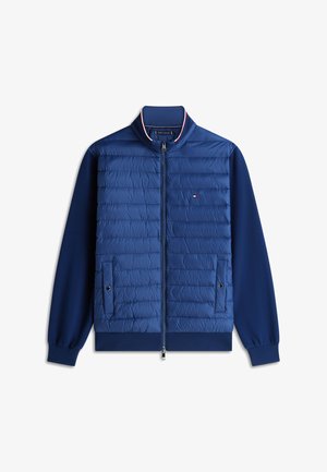 Jachetă albastră matlasată cu fermoar frontal, două buzunare cu capse, manșete și tiv striate și mic logo roșu, alb și albastru pe piept.