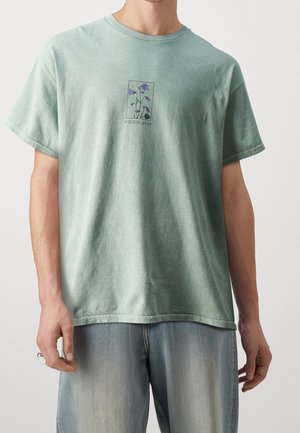 Hombre vestido con una camiseta verde desgastada que tiene una pequeña ilustración de una flor silvestre morada y el texto "PLANTAS SILVESTRES," combinada con unos jeans de mezclilla azul claro.