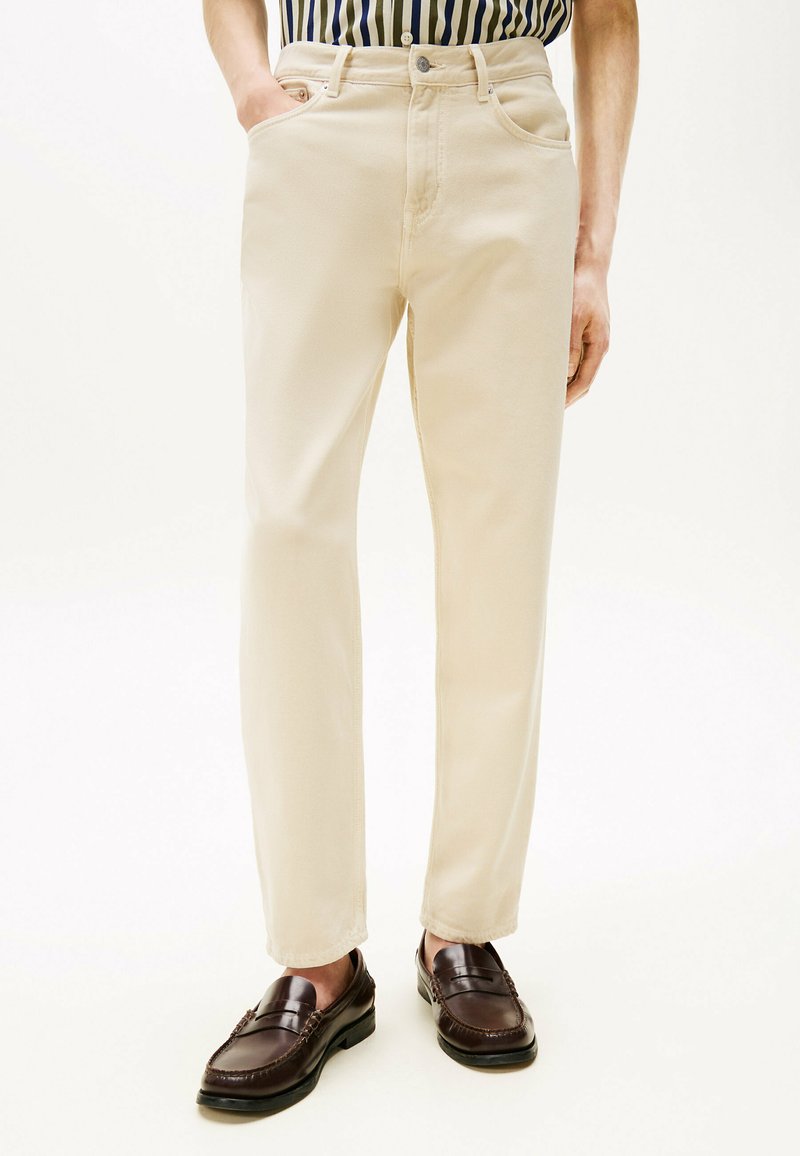Beige Baumwollhose mit geradem Bein, ausgestattet mit Fronttaschen und einem Knopfverschluss, kombiniert mit dunkelbraunen Slippern.