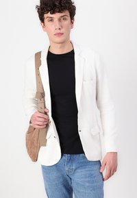 Fehér vászon zakó, amelyet kialakított hajtókával viselnek, fekete pólóval, világoskék farmernadrággal és barna velúr keresztpántos táskával párosítva.