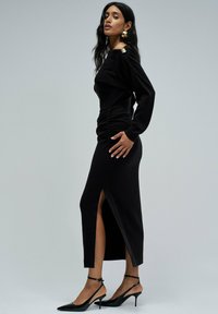 Vestido negro ajustado con mangas largas, fruncido en la cintura y una abertura lateral. Combinado con tacones negros de punta y correas en los tobillos.