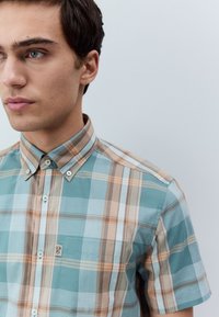 Camicia a maniche corte a quadri nei toni turchese, crema e marrone. Presenta colletto con bottoni, tasca e trame a contrasto nel tessuto.