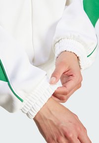 Valkoinen pommikoneen takki vihreillä yksityiskohdilla, jossa on poimutetut hihansuut. Kangas näyttää sileältä, ja hihassa on vetoketju ja joustava yksityiskohta.