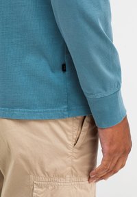 Langärmliges, türkisfarbenes Shirt aus weichem Stoff, mit Rundhalsausschnitt und einem kleinen Markenetikett an der Seite, neben beigefarbenen Shorts mit einer Seitentasche.