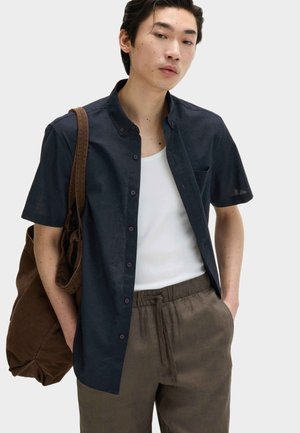 Ung mand iført en marineblå kortærmet skjorte med knapper over en hvid tanktop, brune bukser med snor i taljen og bærer en brun taske på den ene skulder.
