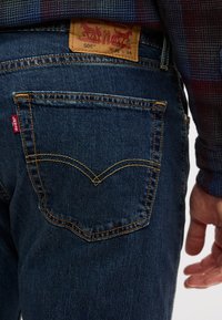 Πίσω όψη ατόμου που φοράει μπλε τζιν Levi’s 505 με εμφανή ραφή στις τσέπες, κόκκινη ετικέτα και δερμάτινη ετικέτα με το λογότυπο στη μέση.