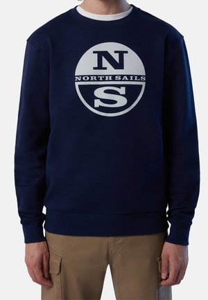 Mężczyzna noszący granatową bluzę z białym logo "NORTH SAILS" oraz beżowe spodnie typu cargo, stojący na jednolitym tle.