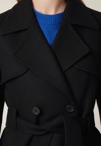 Czarny dwurzędowy płaszcz z szerokimi klapami, z tkaninowym paskiem i kontrastowym niebieskim swetrem pod spodem. Widać dwa czarne guziki.