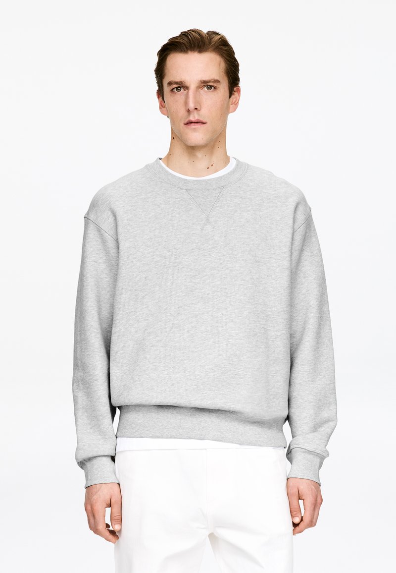 Jeune homme aux cheveux bruns courts portant un sweat-shirt gris clair sur une chemise blanche et un pantalon blanc, debout devant un fond blanc uni.