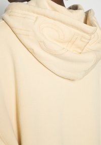 Beige huvtröja med en mjuk textur, som har en stor präglad logotyp på huvan, lös passform och minimalistisk design.