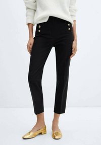 Pantalon noir sur mesure taille haute, avec des détails de boutons dorés à l'avant. Tissu lisse et coupe slim. Associé à des ballerines dorées.