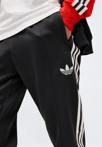Άτομο που φοράει μαύρο αθλητικό παντελόνι Adidas με λευκές ρίγες και λογότυπο, λευκό πουκάμισο και κόκκινο μακρυμάνικο με μαύρες και λευκές ρίγες.