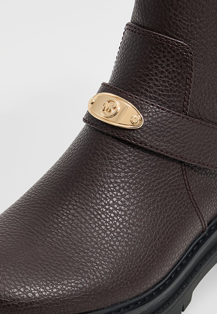 Botín de cuero marrón oscuro con acabado texturizado y una correa de hardware dorado con un emblema de logo. Suela de goma para tracción.