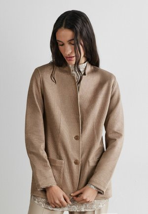 Femme aux longs cheveux noirs portant une veste beige boutonnée par-dessus un haut à motifs, regardant vers le bas avec les mains près de l'ourlet de la veste.