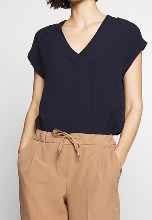Marineblaue V-Ausschnitt-Bluse mit kurzen Ärmeln, im lässigen Schnitt. Kombiniert mit hellbraunen, Hosenträgerhosen mit Taschen und einem Zugband. 