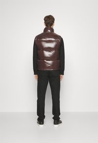 Braune glänzende Steppweste mit hohem Kragen, oversized Design und horizontalen Nähten, getragen über einem schwarzen Langarmshirt und einer Hose.